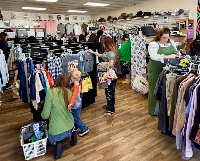 A busy retail clothing store interior with multiple people browsing racks of garments on a wood floor. Shoppers are seen examining clothes, and clothing displays line the aisles under overhead fluorescent lighting.