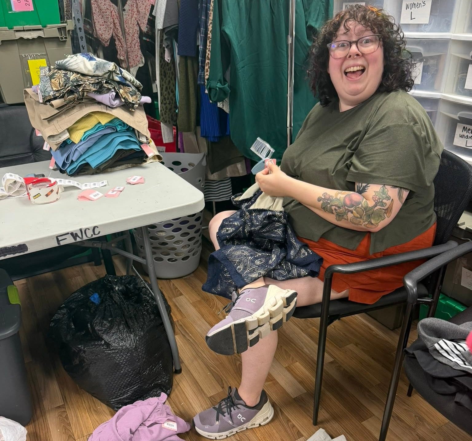 A person sitting in a chair in a cluttered clothing storage or resale room, holding a phone and smiling. The room contains racks of clothes, a folding table covered in garments, a black trash bag, and stacks of folded clothes on the wooden floor.